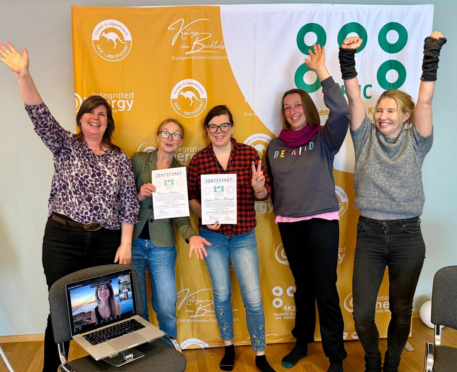 Fünf Frauen vor dem AKJC-Banner, drei jubeln mit erhobenen Armen, zwei halten ihr Zertifikat «Systemischer Familiencoach», vorn ein Laptop mit einer Online-Teilnehmerin